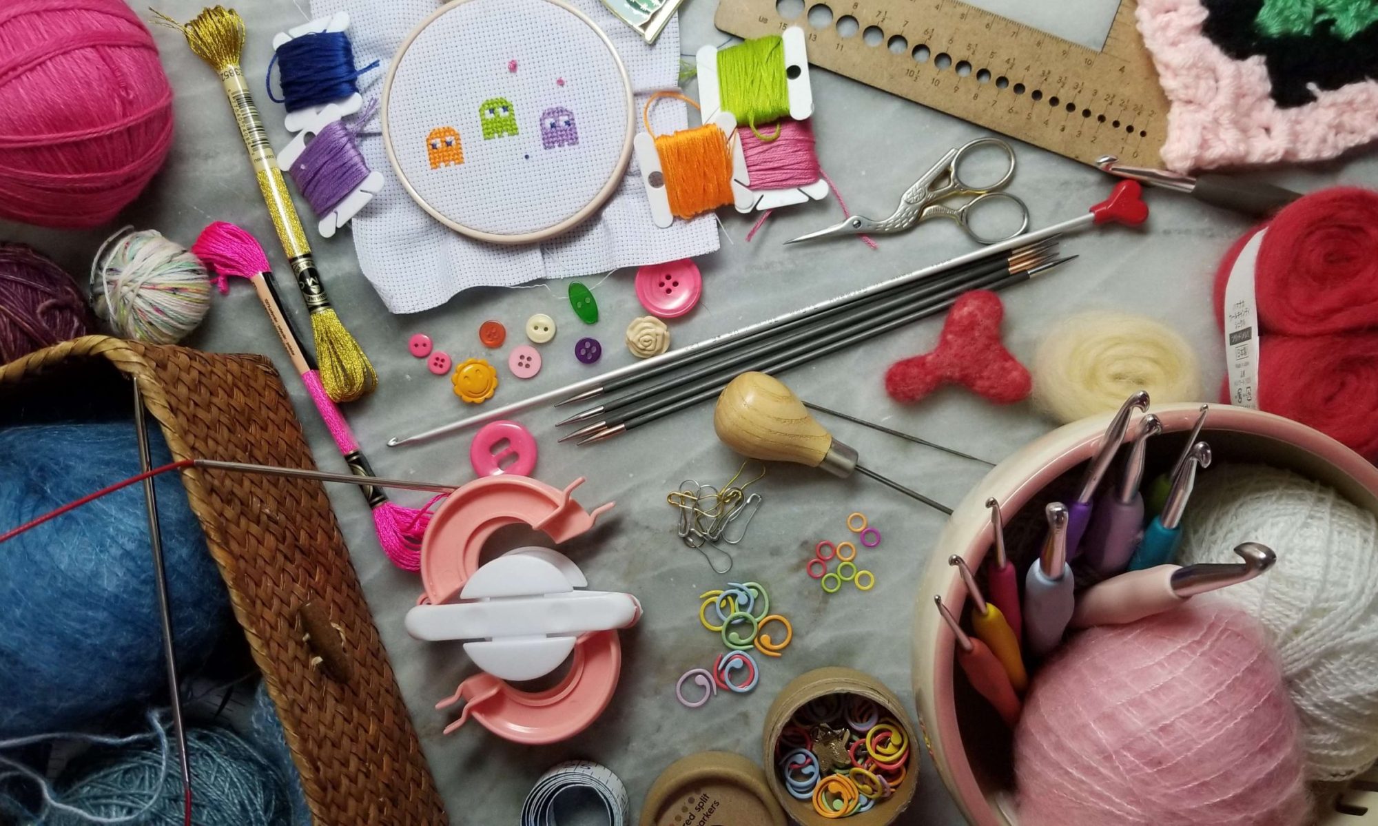 Craft supplies for cross stitch, knitting, crochet, needle felting and notions laid out on a marble countertop background.