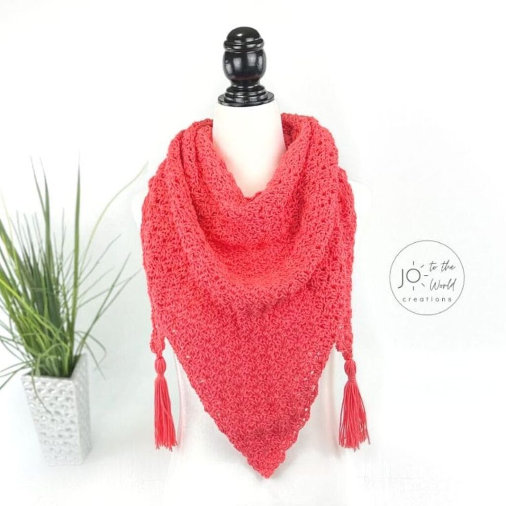 A coral-coloured lace crochet shawl with tassels on either corner wrapped around a mannequin bust.