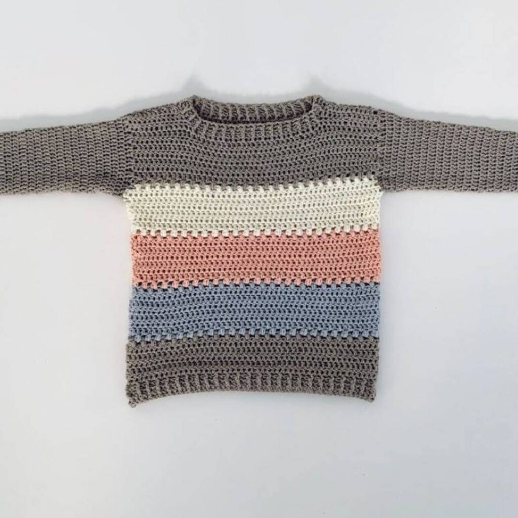 Crochet sweater laid flat on a white background. It is grey, with three thick stripes along the body made of white, pink, and blue.