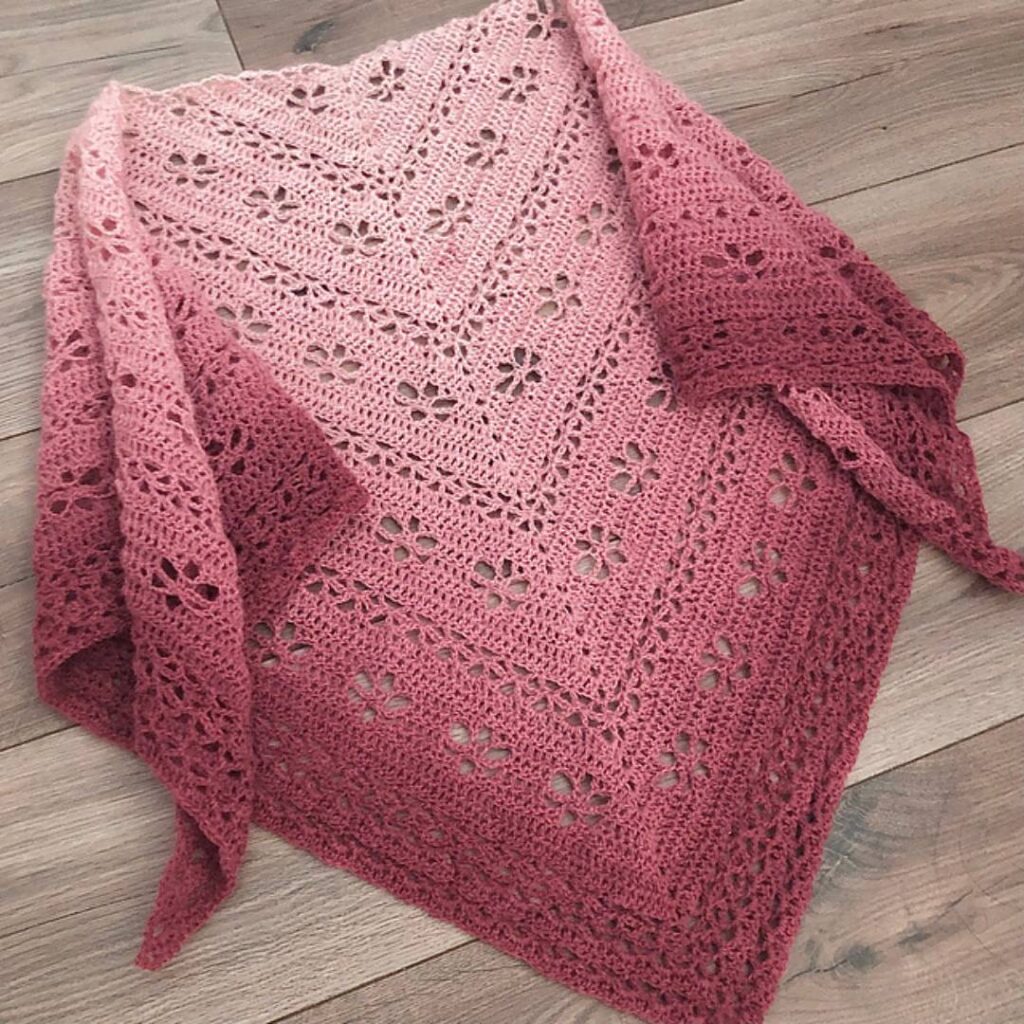 Lacy crochet triangle shawl laid out flat. It is an ombre from light pink at the starting point to a dark maroon at the edges.