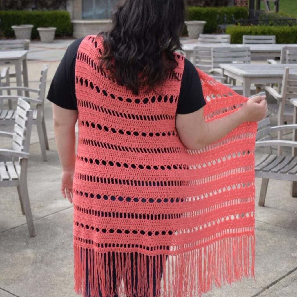 Person facing away from camera wearing a long bright coral lacy cardigan without sleeves. The cardigan has fringe along the bottom hem.