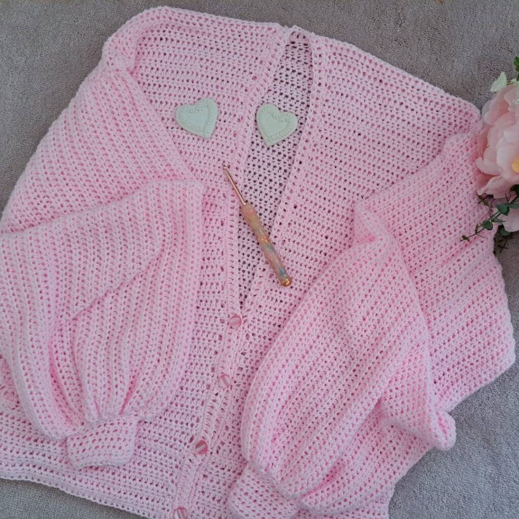 Pink crochet cardigan with airy fabric and balloon sleeves laid out flat on a grey background.