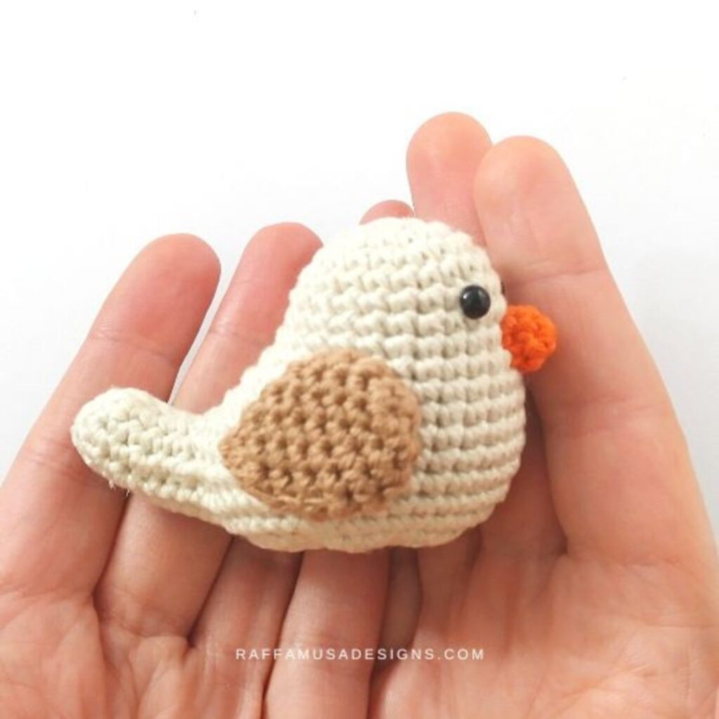 Small crocheted bird laying on its side in someone's hand. The bird is a soft beige, its wing is a slightly darker brown.