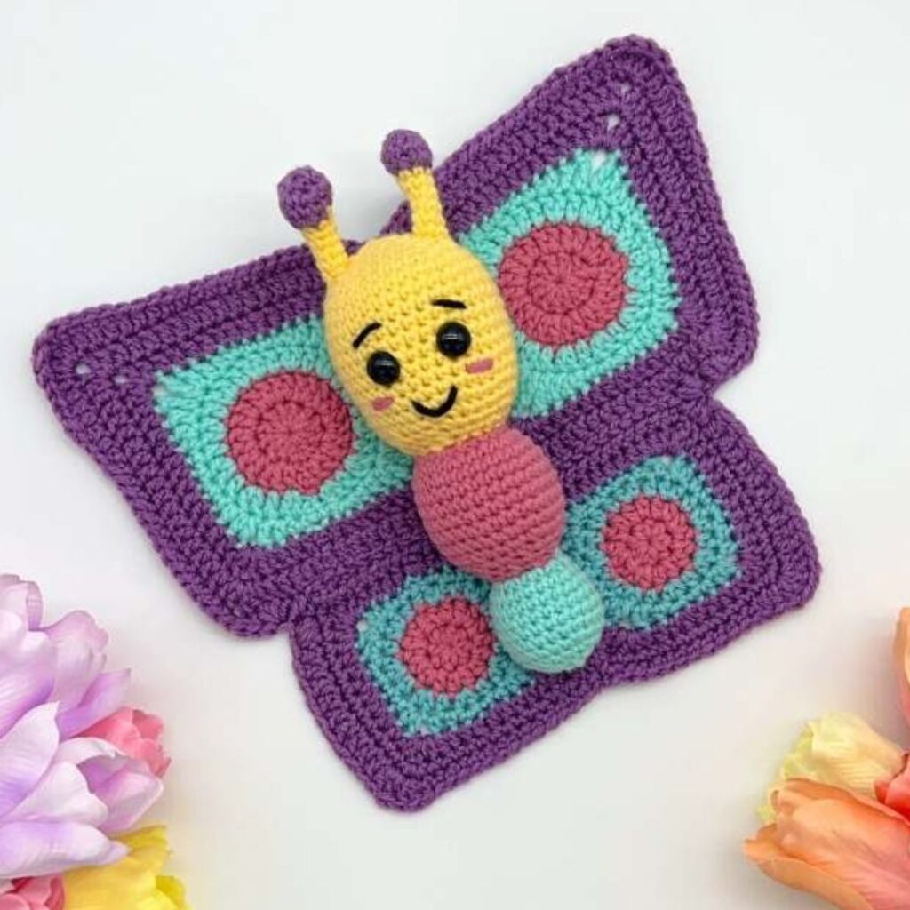 Crochet butterfly with a plush body and large, flat wings. It has an embroidered face. 