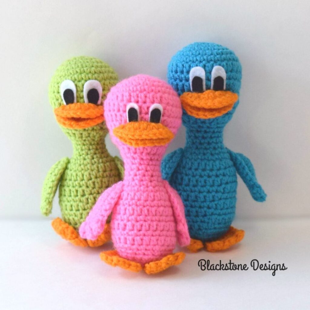 Three crochet ducklings in a row. From left to right they are green, pink, and blue.