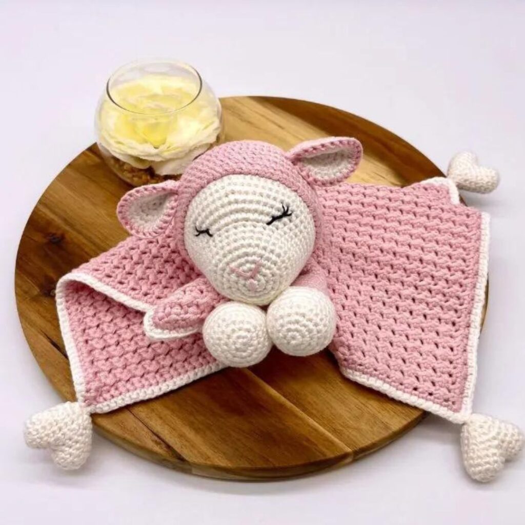 A small pink baby blanket with a lamb head crocheted in the center. There are white hearts on each corner of the blanket.