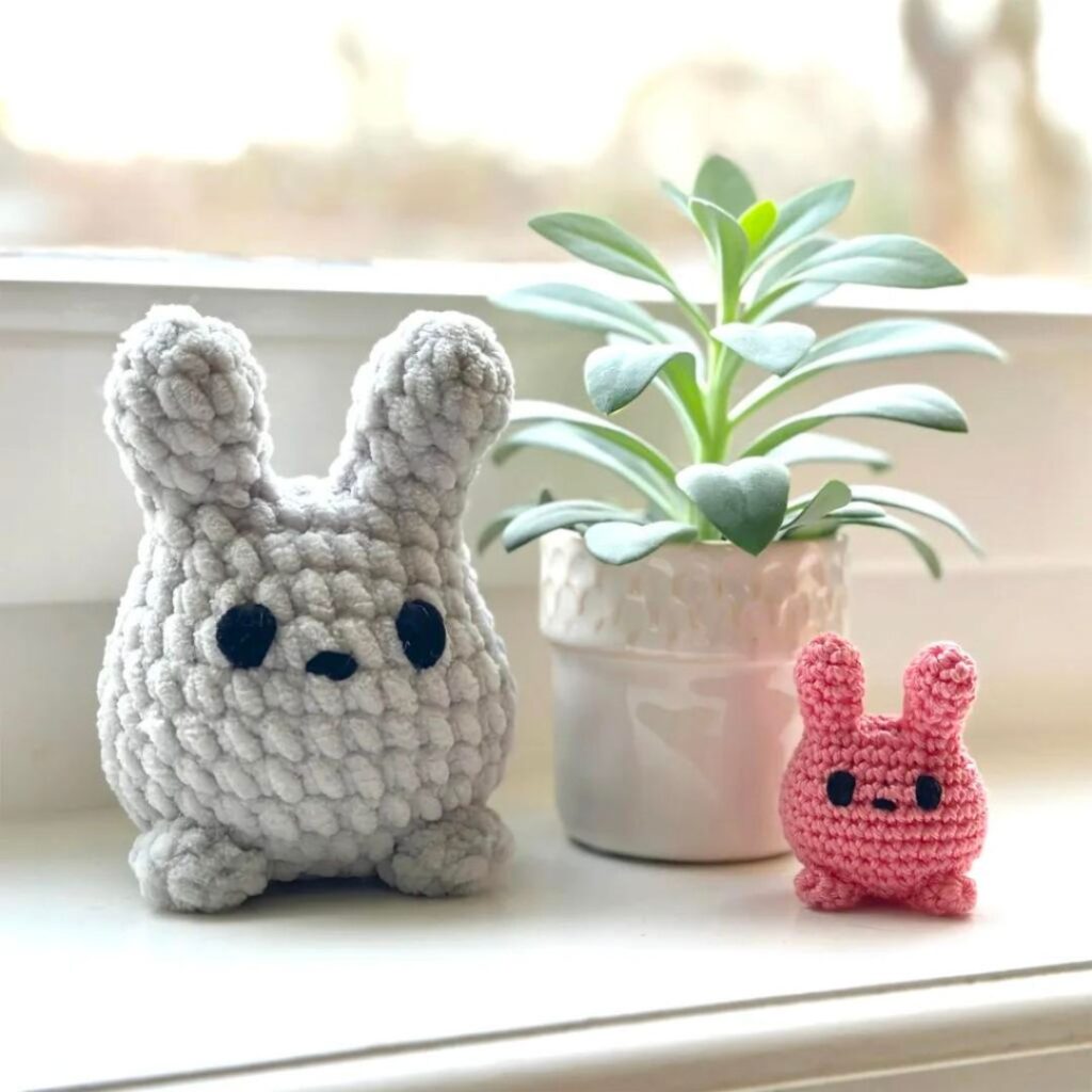 Two crochet bunnies in front of a potted plant. The left bunny is large and made of a soft yarn. The right bunny is keychain sized.